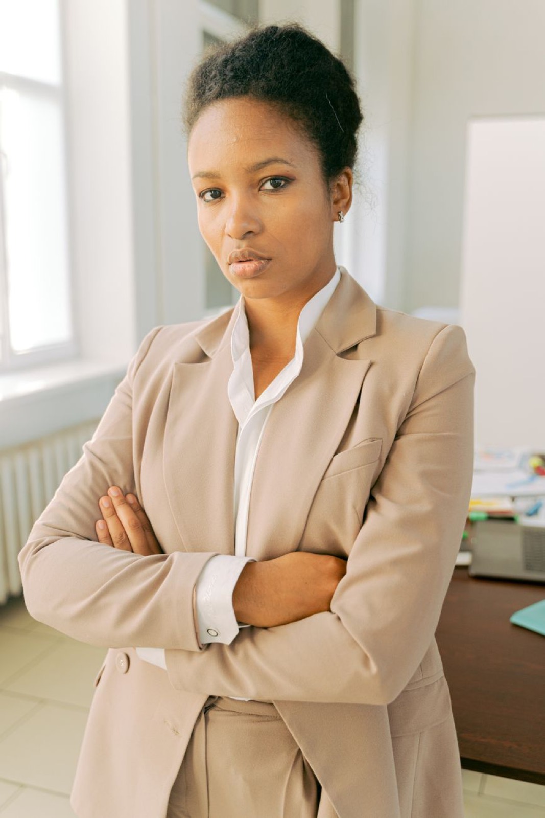 A woman in a camel blazer, arms folded, by a window